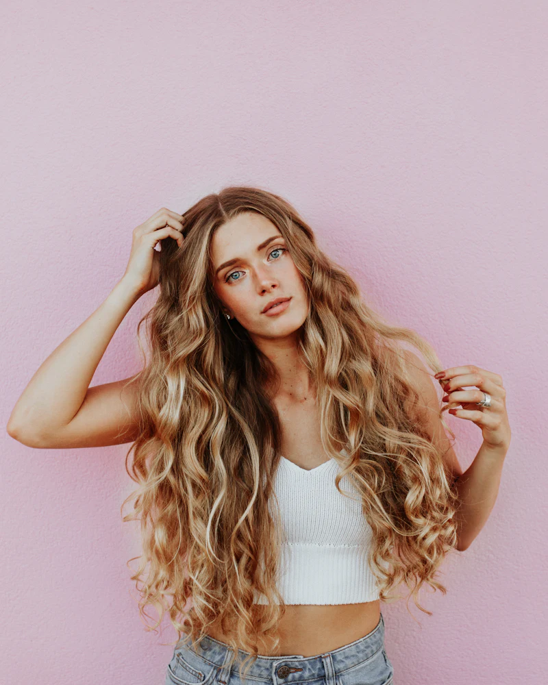 Woman with long blonde waves and darker roots standing against a pink wall